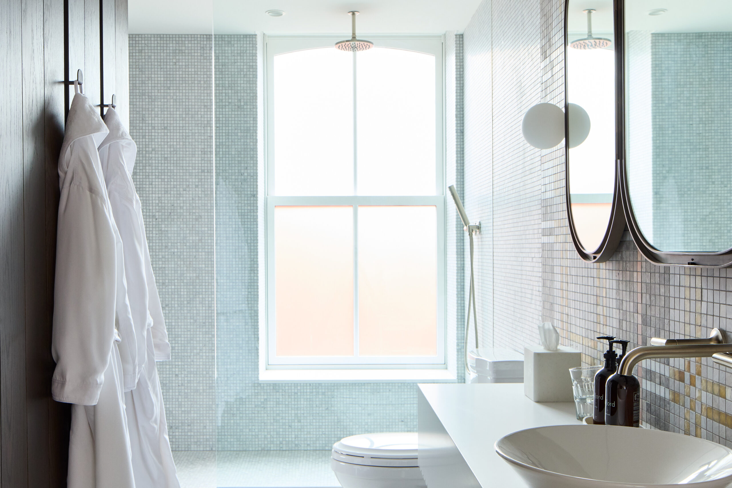 Brightly lit bathroom tiled from floor to ceiling with two white robes