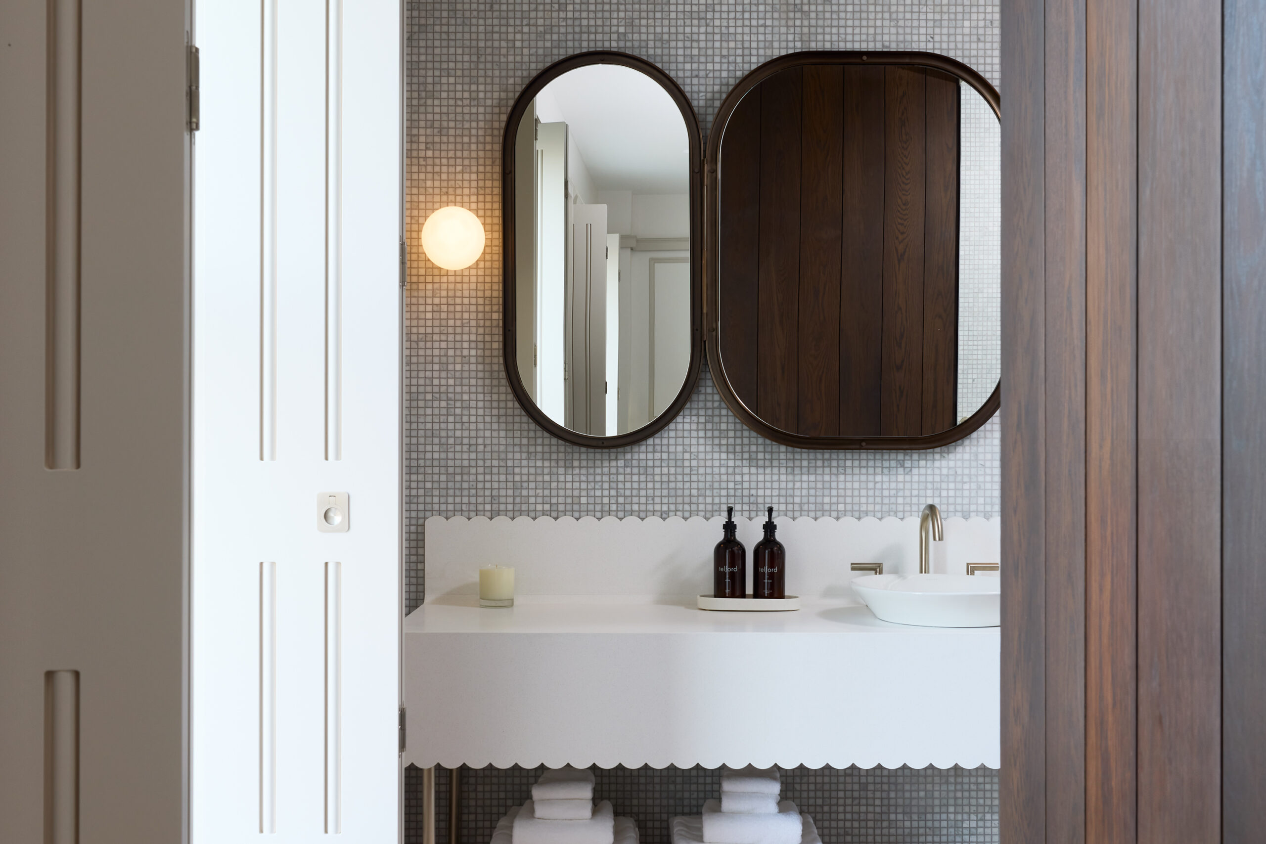 Black rimmed mirrors on a tiled wall, white counter, and white bowl sink with gold fixtures