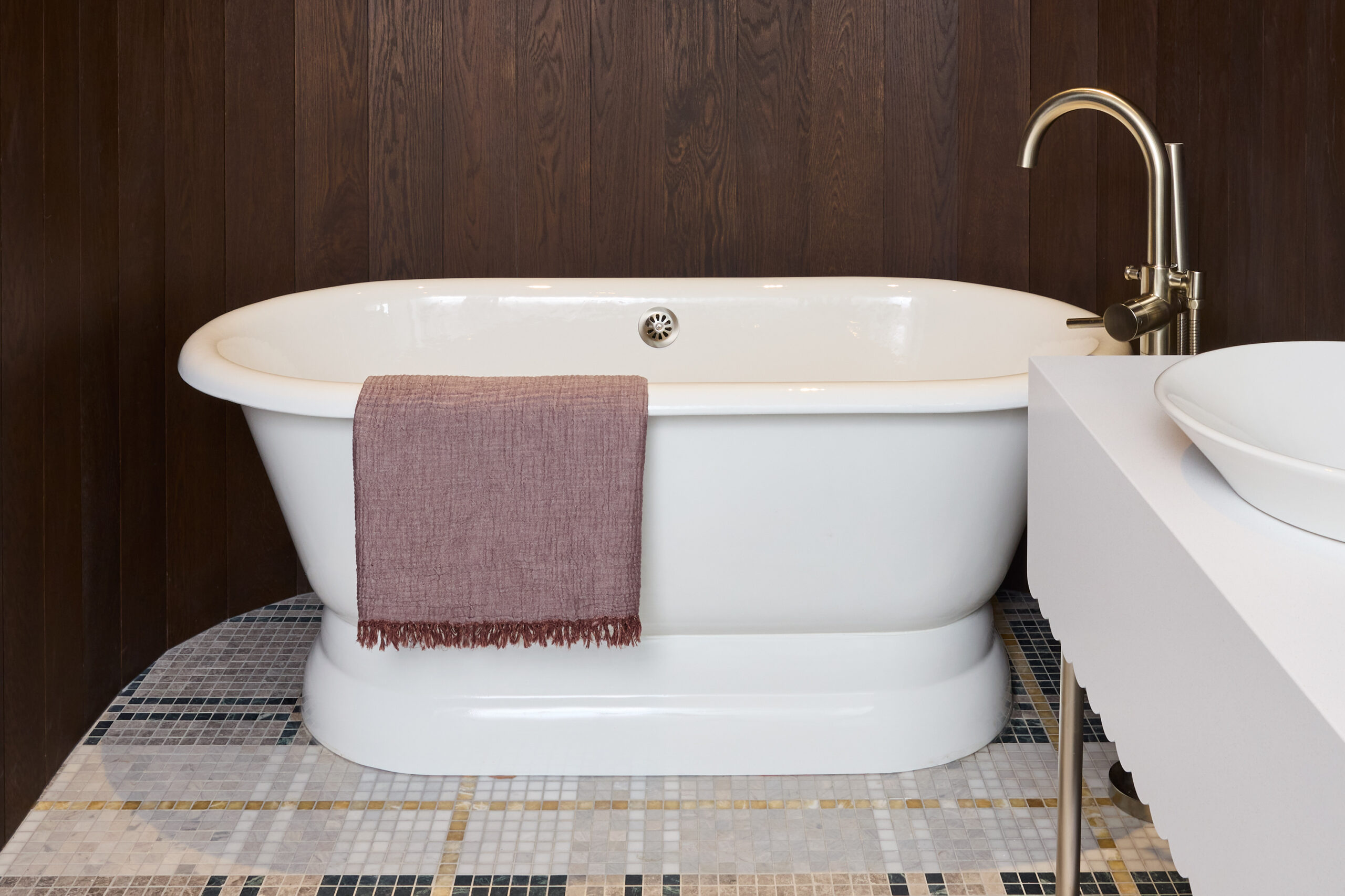 White stand alone tub, on a dark wood panelled background and gold fixtures