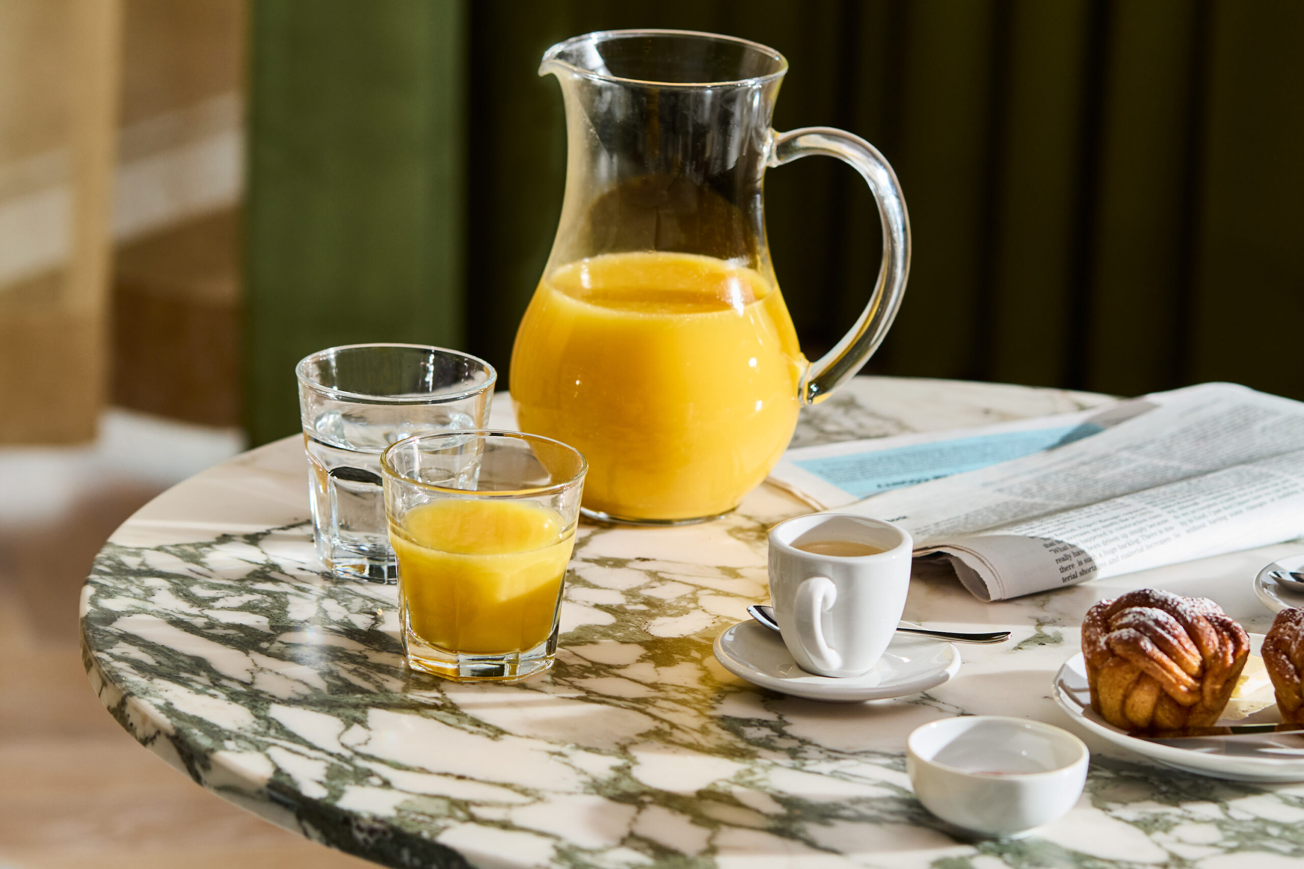 Jug of orange juice with a glass half full of orange juice and two muffins on a side table